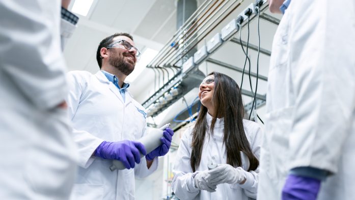 4 people wearing lab coats stand in a room talking and smiling.